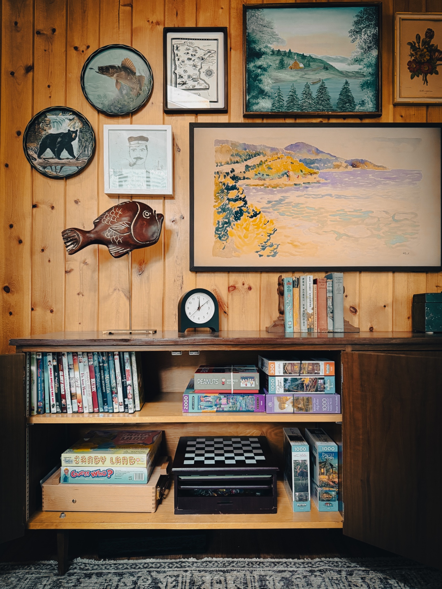 Cabinet stocked with books, games, and puzzles beneath the cabin art wall